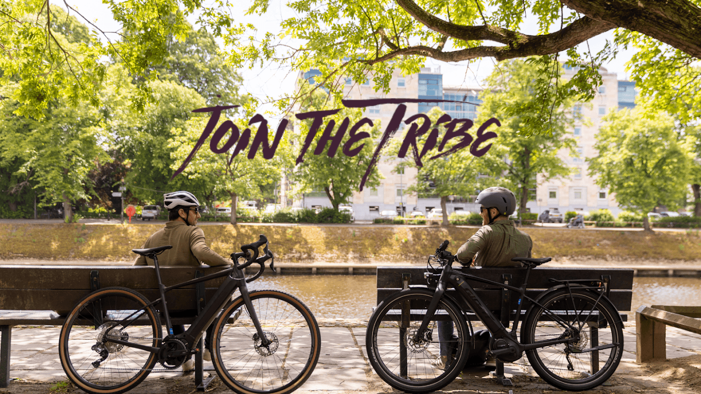 Two cyclists sit on benches by a river, surrounded by trees, with "JOIN THE TRIBE" text above them.