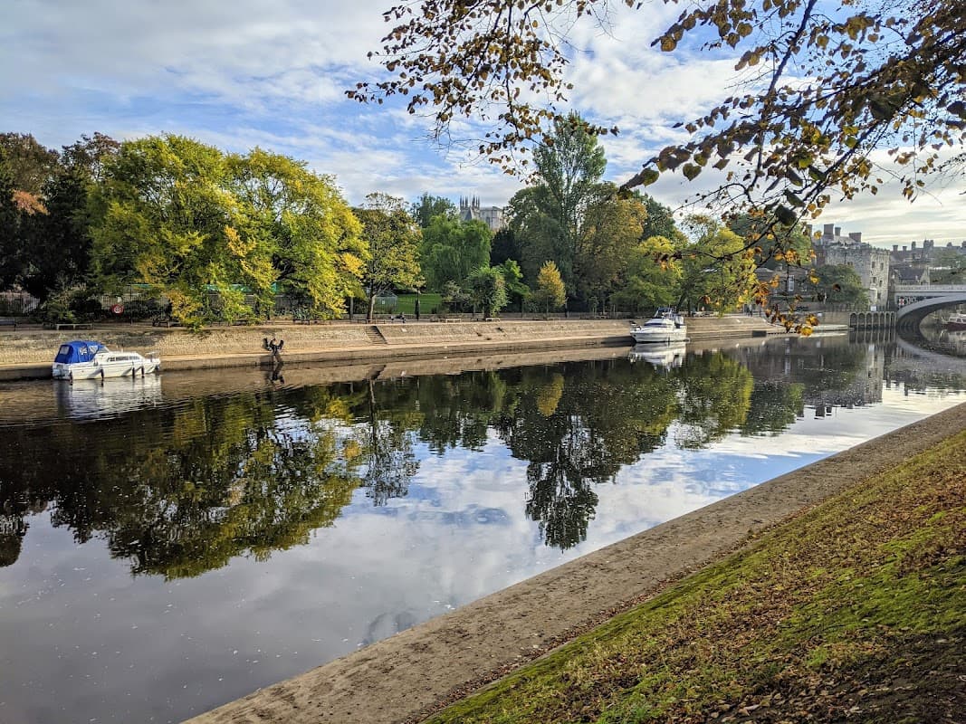Pay & Display car park by a river, with trees and boats reflecting on the water under a blue sky.