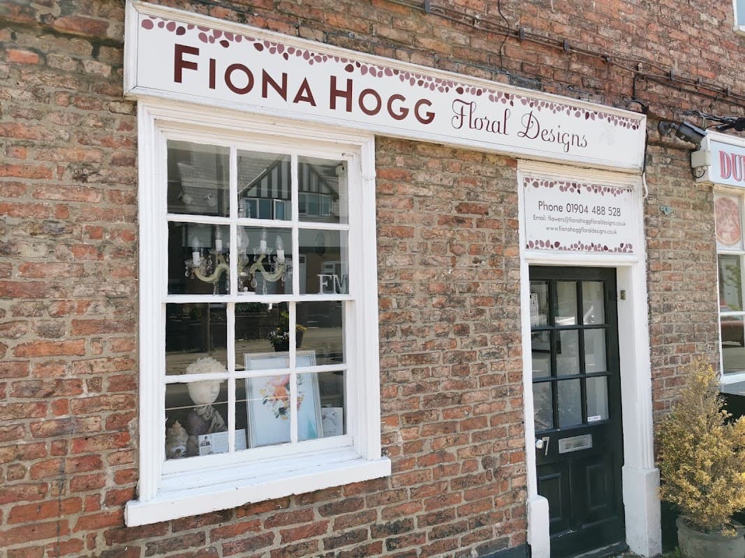 Sign for Fiona Hogg Floral Designs on a brick building, with a window displaying floral arrangements and a door.