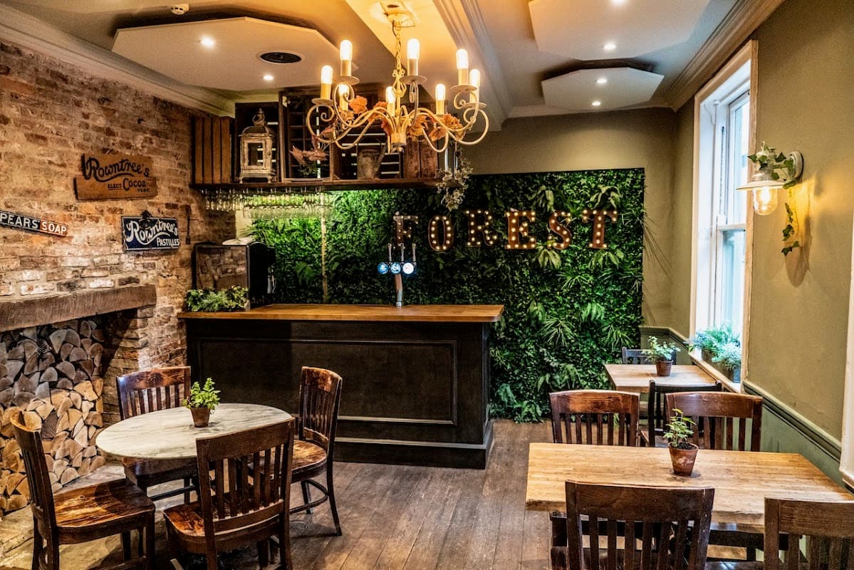 Cozy restaurant interior with a wooden bar, green foliage, rustic decor, and wooden tables and chairs.