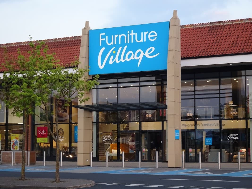 Bright blue sign reading "Furniture Village" on a modern storefront with large glass windows and a tree in front.