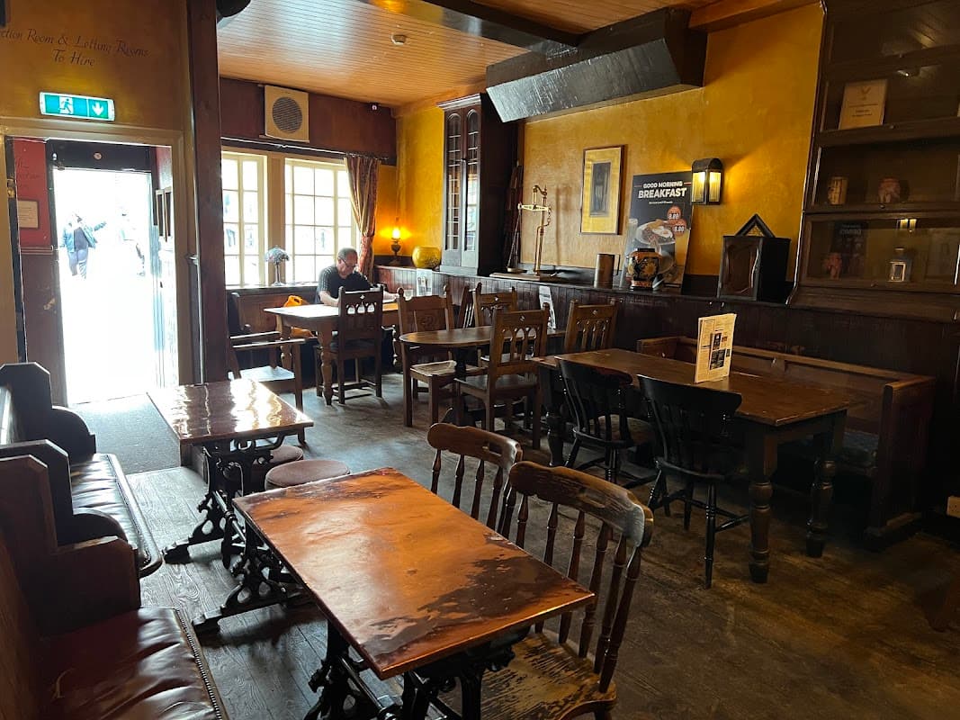 Cozy pub interior with wooden tables, chairs, and warm yellow walls; a lone patron sits at a table near the entrance.
