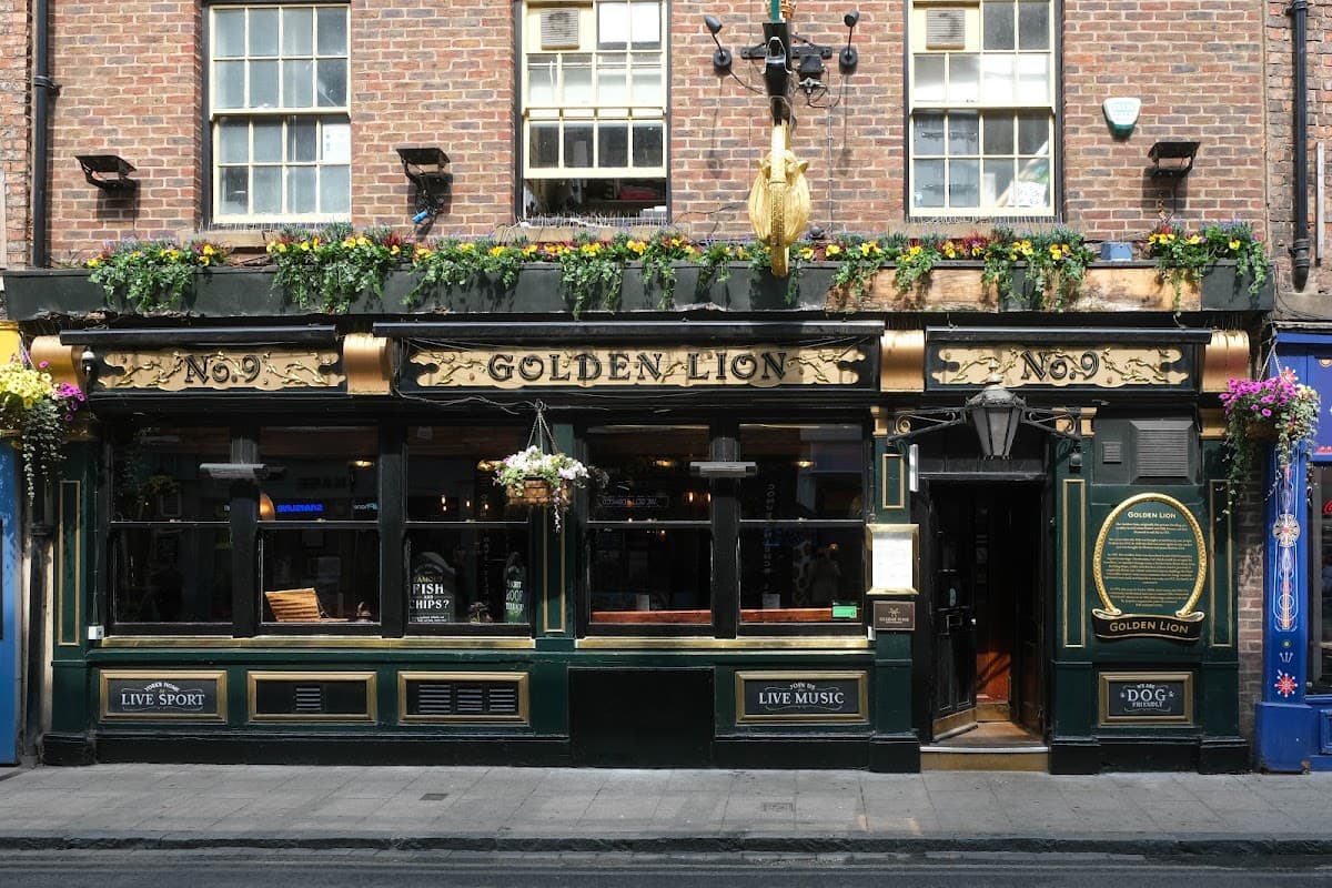 Historic pub with a gold lion sign, floral decorations, and large windows, located on a brick street in York.