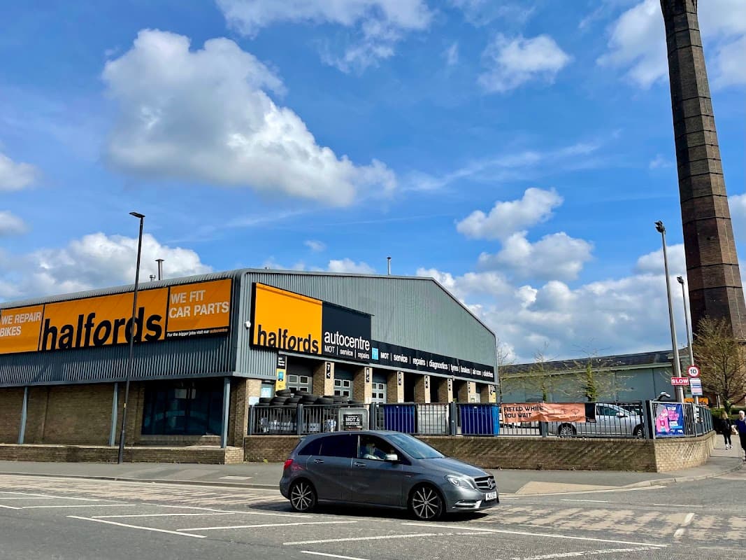 Halfords store with bright signage, cars parked outside, and a tall industrial chimney in the background under a cloudy sky.