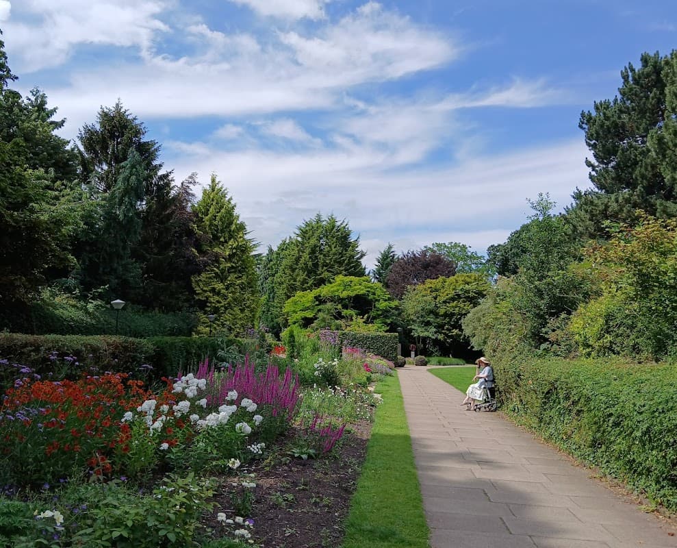 Lush greenery and colorful flower beds line a pathway; a person sits on a bench under a blue sky with fluffy clouds.