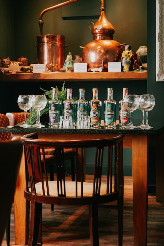 A table with various bottles of Hooting Owl gin, crystal glasses, and copper distilling equipment in a cozy distillery setting.
