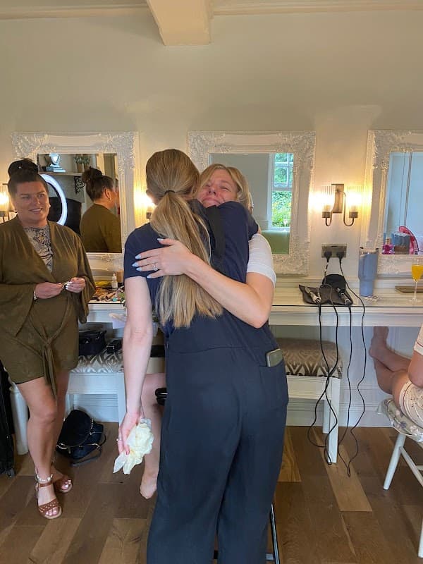 Two women embrace joyfully in a bright room with mirrors, while another woman watches nearby.