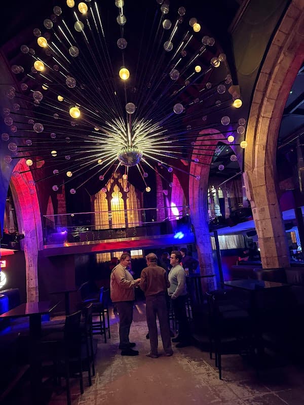 A stylish pub interior with arched stone walls, a large chandelier, and patrons conversing in a vibrant atmosphere.