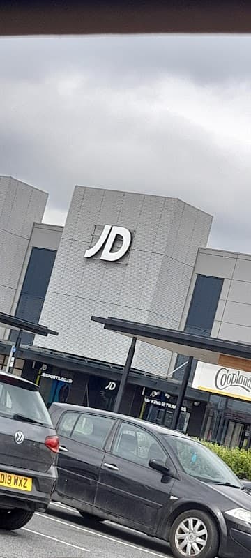 JD Sports store front with a large "JD" sign, surrounded by parked cars and a cloudy sky.