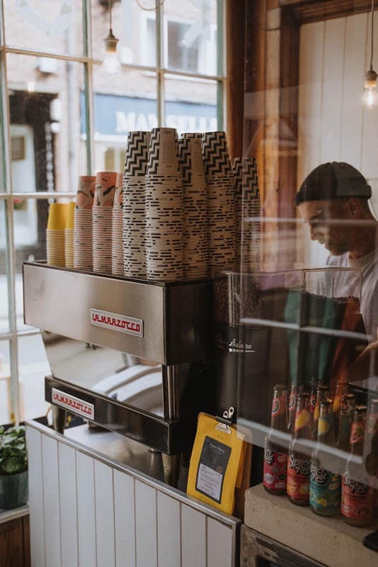 Kiosk Cafe interior featuring a La Marzocco coffee machine, stacked cups, and colorful beverage cans.