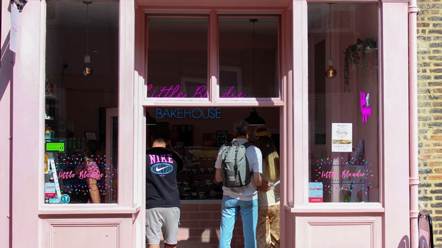 Pink storefront of Little Blondie Bakehouse with customers browsing inside, displaying baked goods and a welcoming atmosphere.