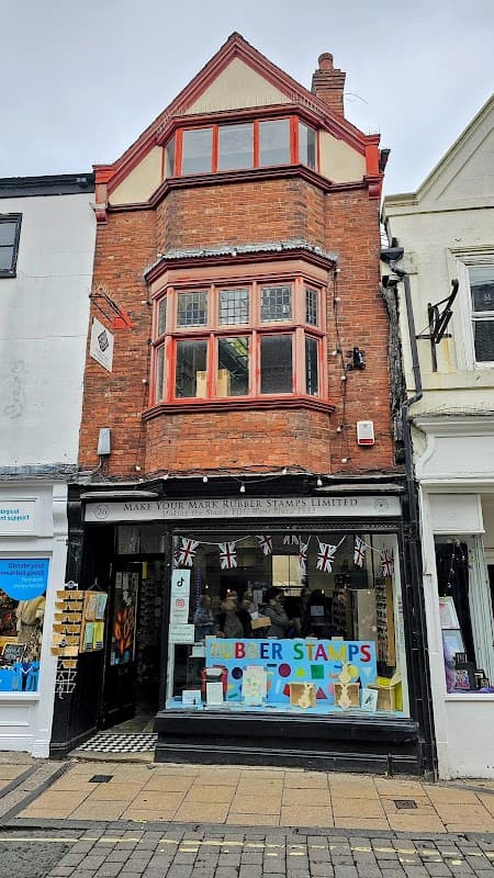 Historic brick shopfront with large windows, featuring "Make Your Mark RUBBER STAMP" sign and colorful display.