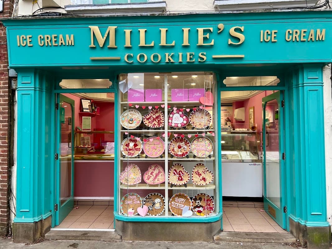 Bright turquoise storefront with "Millie's Cookies" sign, displaying colorful cookie trays and ice cream offerings.