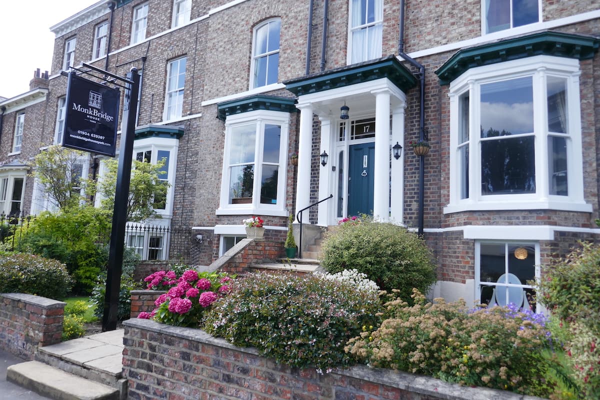 Charming brick facade of MonkBridge Guest House with large windows, colorful flower beds, and a welcoming sign.
