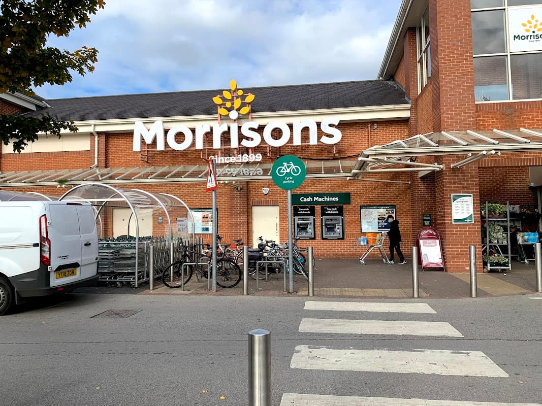 Morrisons grocery store entrance with bike racks, cash machines, and shopping carts outside.