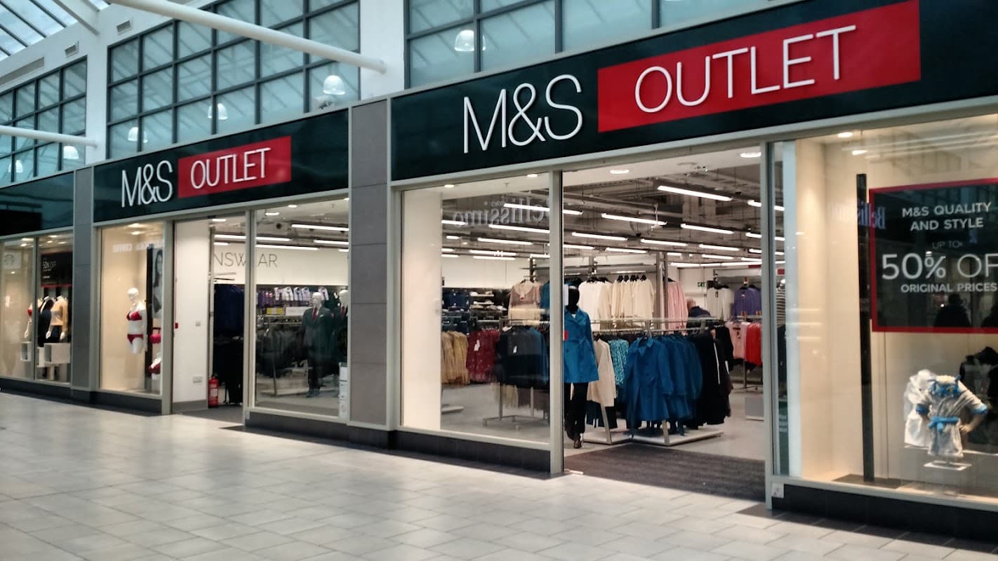 M&S Outlet storefront with large signs, displaying various clothing items and a sale banner for 50% off.