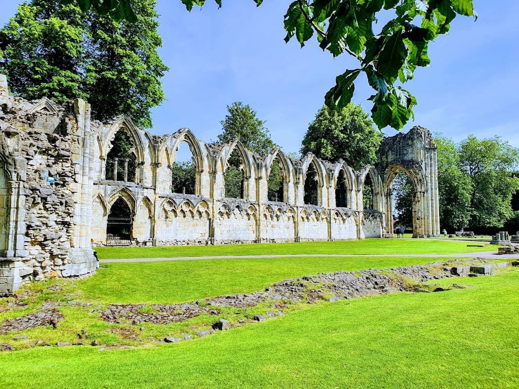 Historic stone ruins with arched windows, surrounded by lush green grass and tall trees under a clear blue sky.