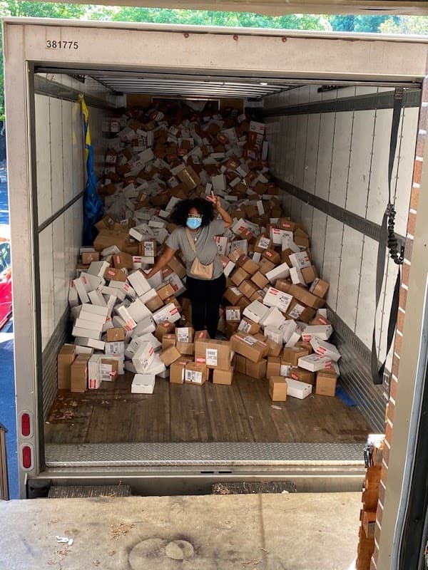 A woman in a mask stands among a large pile of cardboard boxes inside a delivery truck.