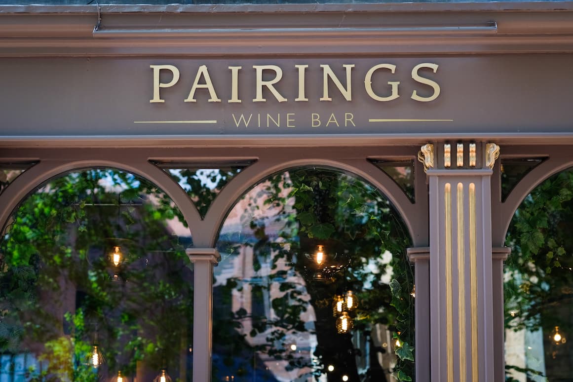 Sign for "Pairings Wine Bar" with elegant lettering and reflections of greenery and lights in the windows.