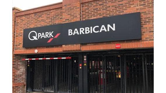 Sign for Q-Park Barbican with a red barrier and brick wall background.