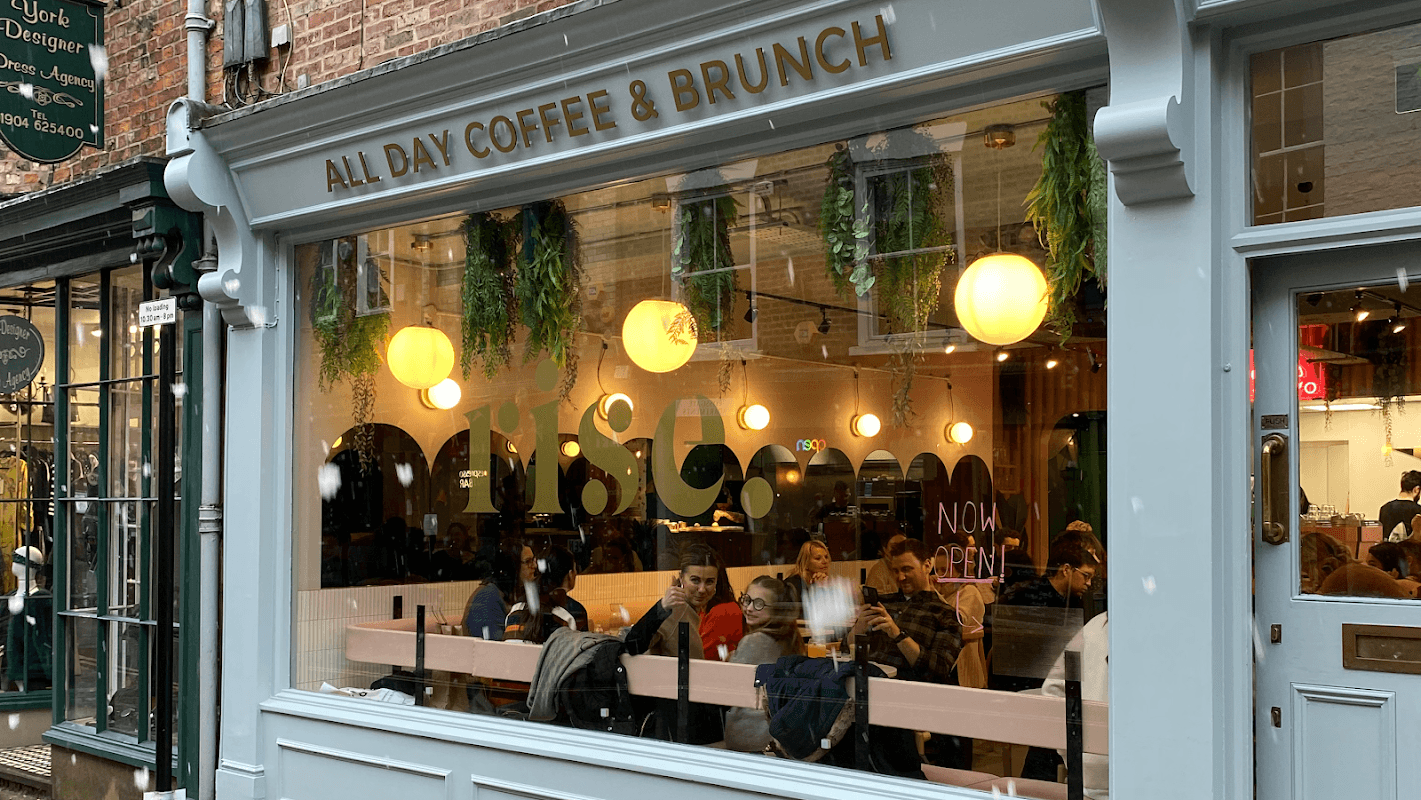 A cozy restaurant with large round lights, indoor plants, and diners enjoying coffee and brunch through a glass window.