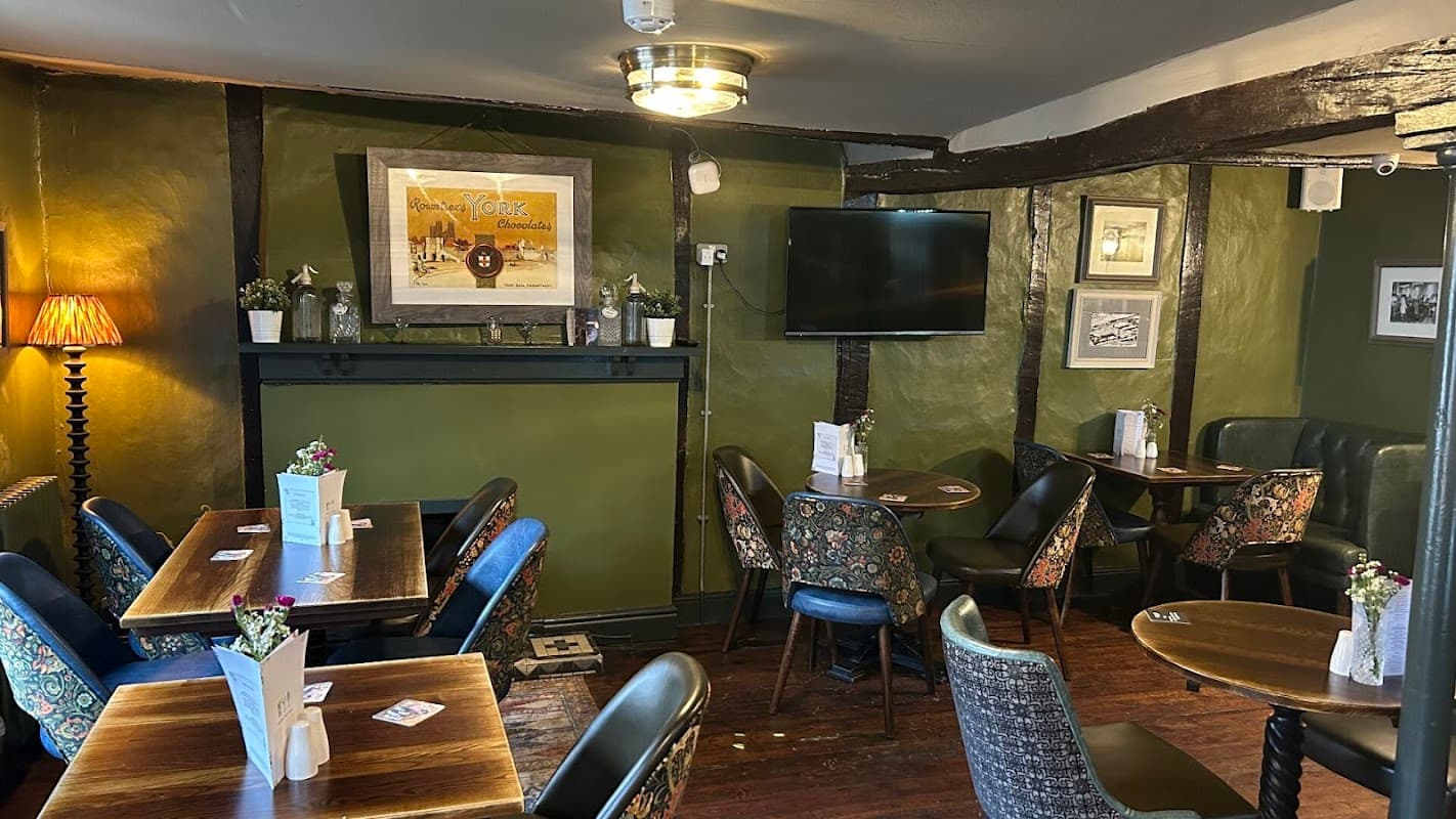 Cozy pub interior with wooden beams, green walls, floral chairs, small tables, and framed artwork.