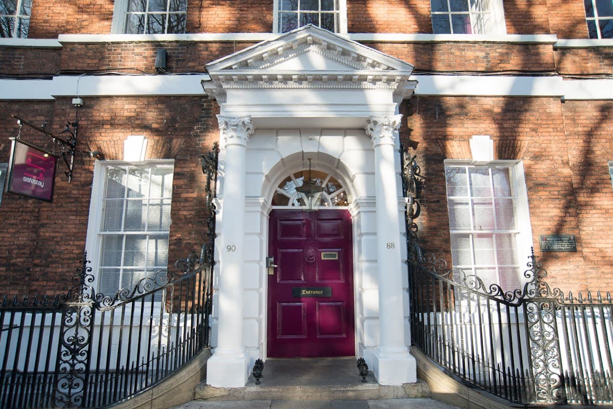 Historic brick building with a grand white entrance and a maroon door, framed by decorative iron railings.