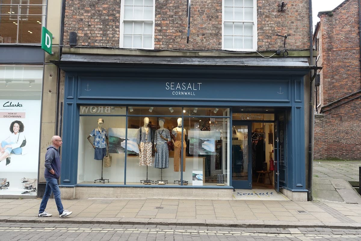 Seasalt Cornwall storefront displaying clothing and accessories, with a pedestrian walking by on a cobblestone street.