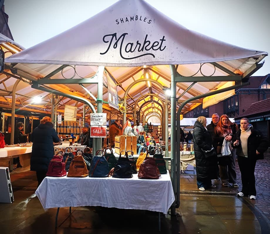 Stalls at Shambles Market display bags and food items, with shoppers browsing in a vibrant, bustling atmosphere.