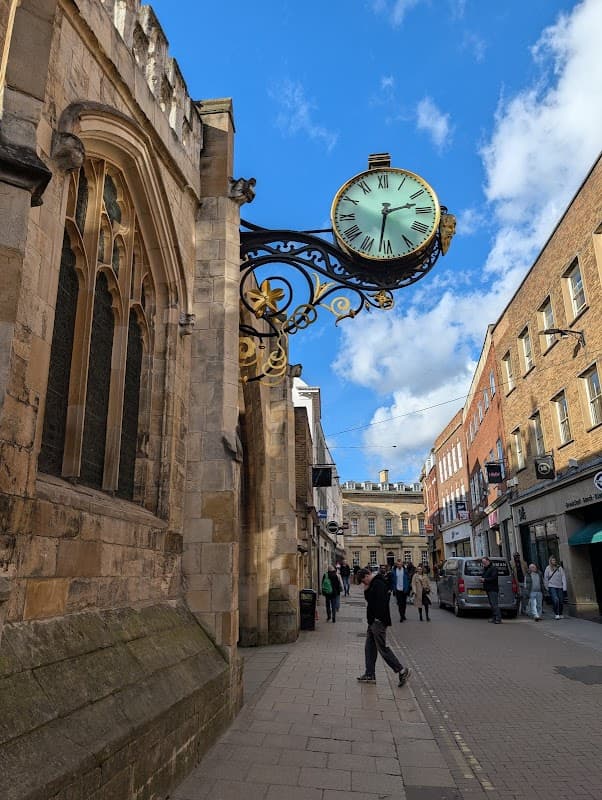 Historic building with ornate clock, cobblestone street, shops, and people walking under a blue sky with clouds.