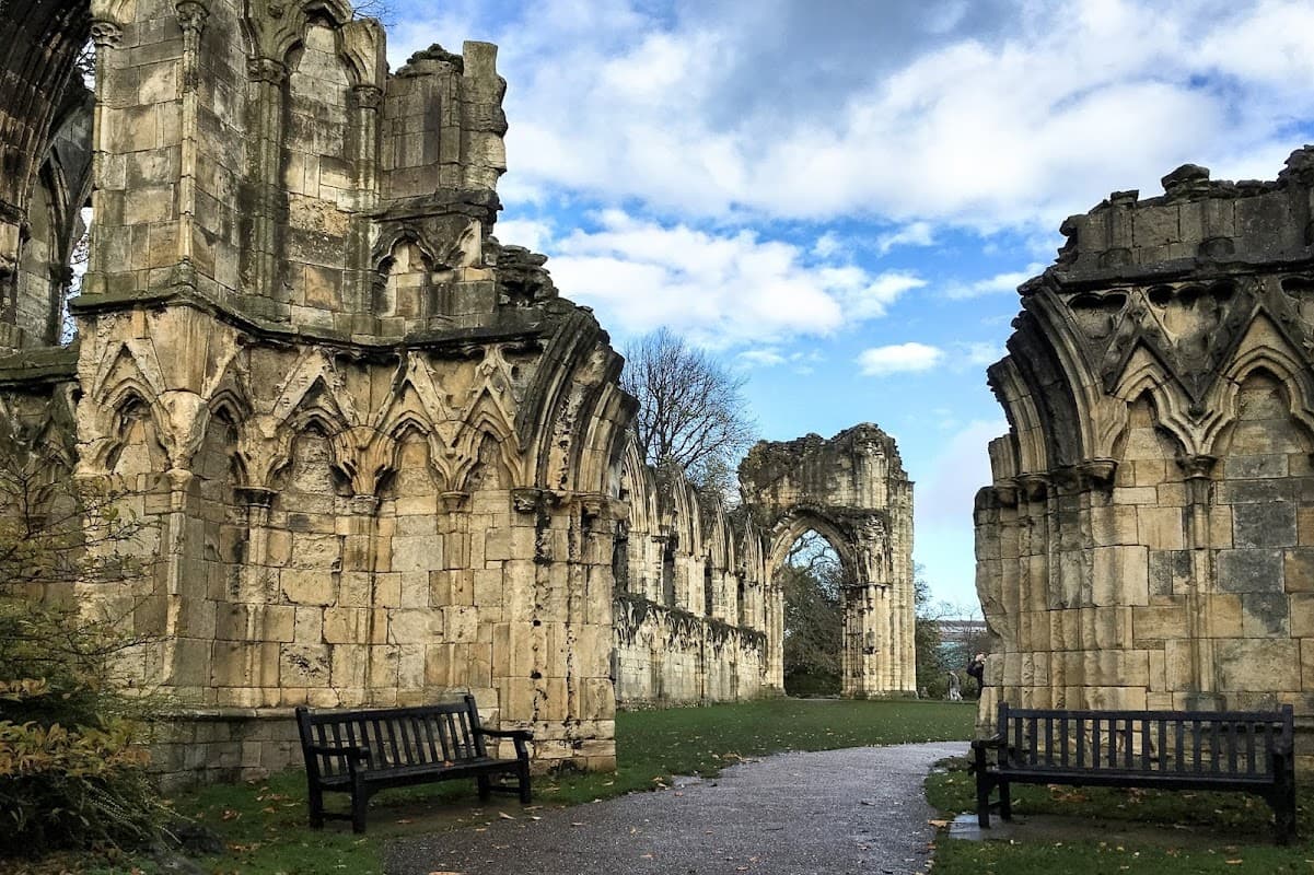 Ruins of St Mary's Abbey with intricate stone arches, benches, and a path under a blue sky with scattered clouds.
