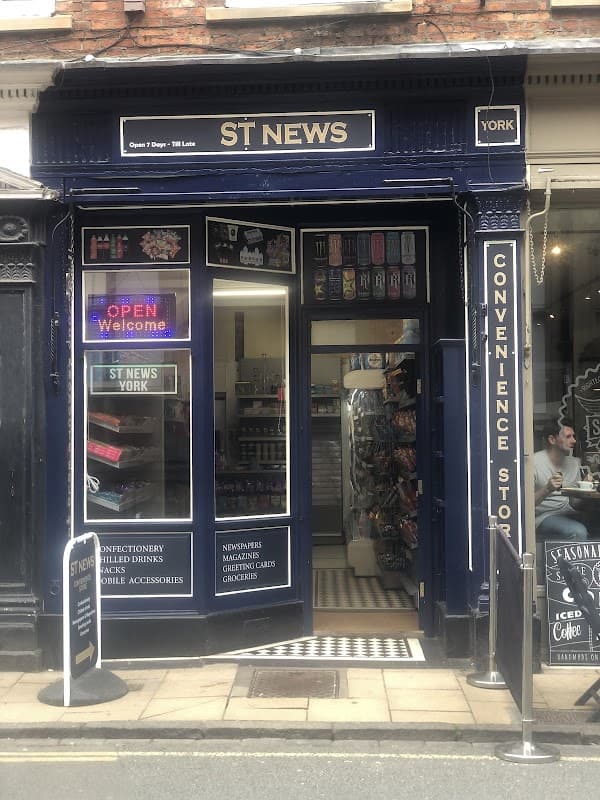 ST NEWS York storefront with open sign, displaying convenience items, snacks, and drinks. Brick building with blue facade.