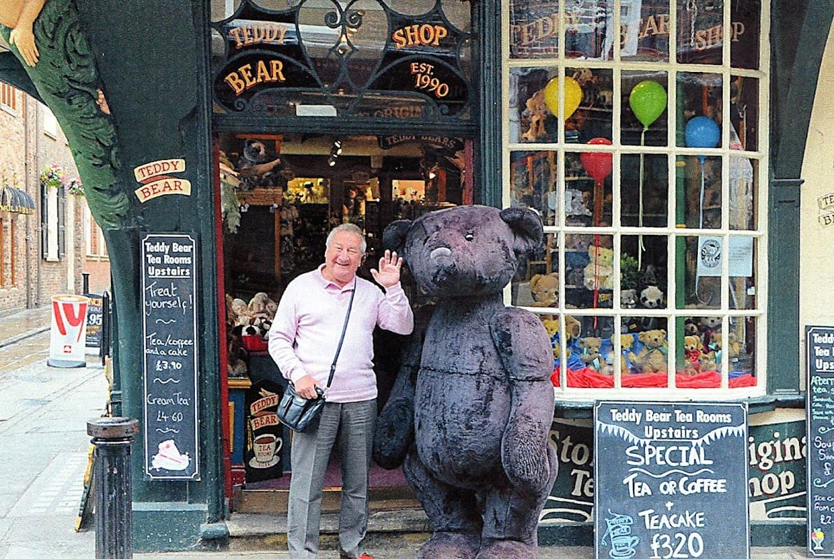 A man waves beside a large teddy bear outside a charming toy shop with colorful balloons and signs in York.