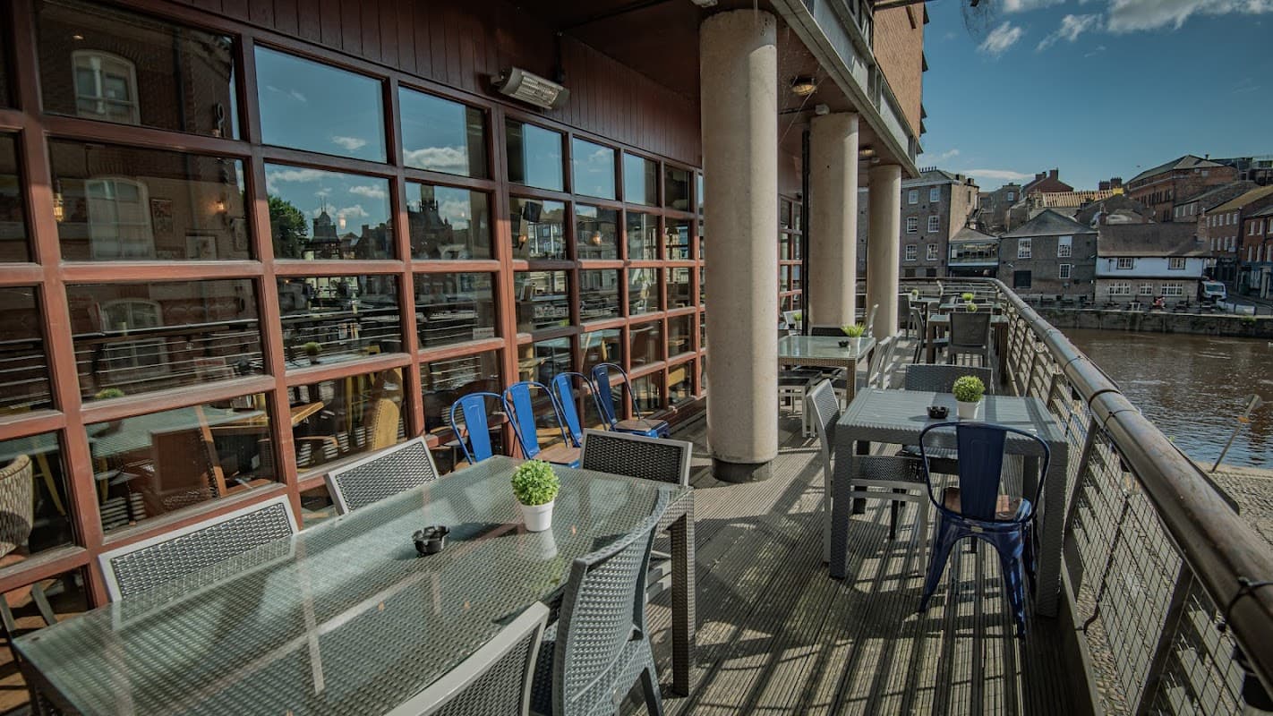 Outdoor seating area with glass tables and chairs overlooking a river in York, Yorkshire. Bright, sunny day.