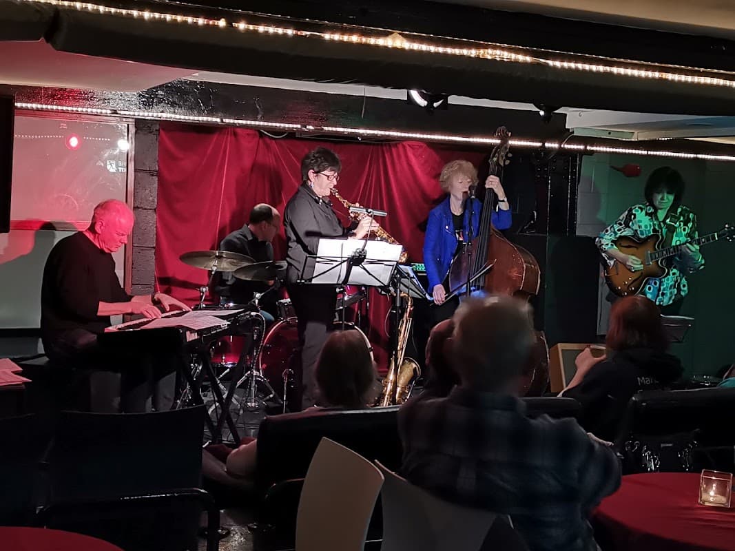 A jazz band performs in a dimly lit basement venue, with musicians on piano, saxophone, bass, and guitar.
