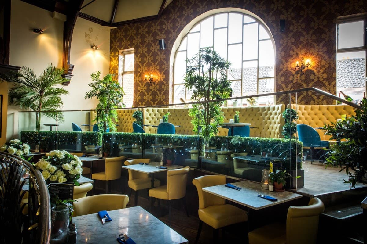 Elegant bar interior featuring plush seating, greenery, and large windows with decorative wallpaper and soft lighting.