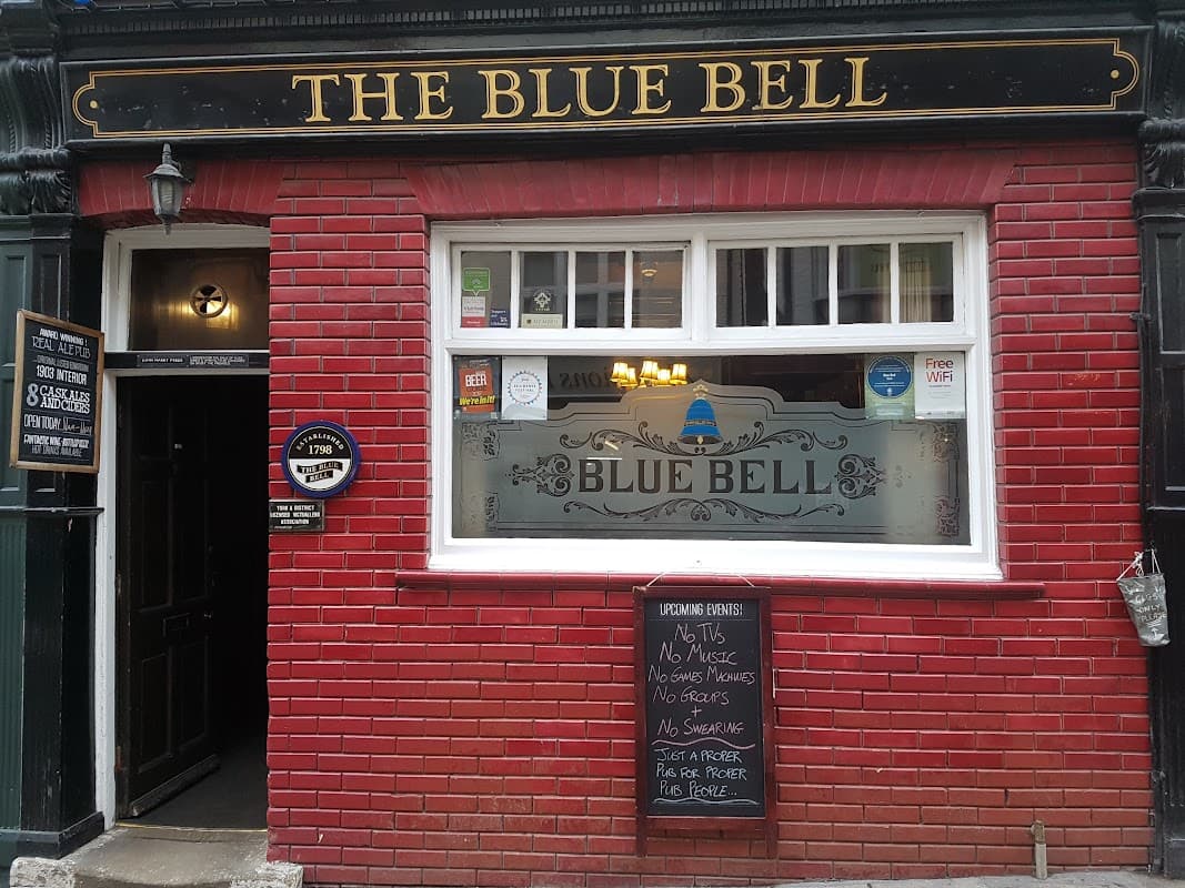 Traditional pub exterior with red brick, large window displaying "The Blue Bell," and a chalkboard menu outside.