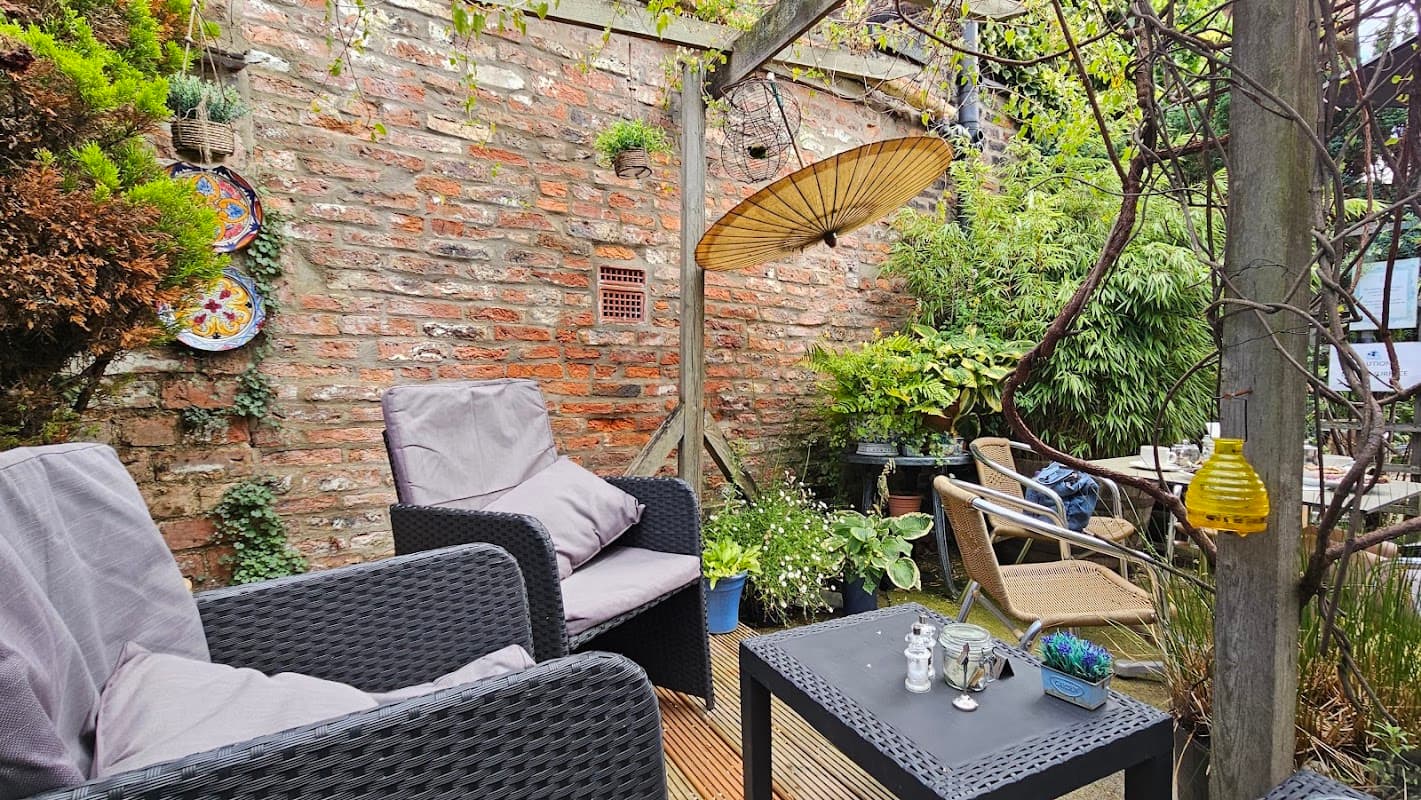 Cozy outdoor seating area with wicker chairs, greenery, and a decorative umbrella against a rustic brick wall.