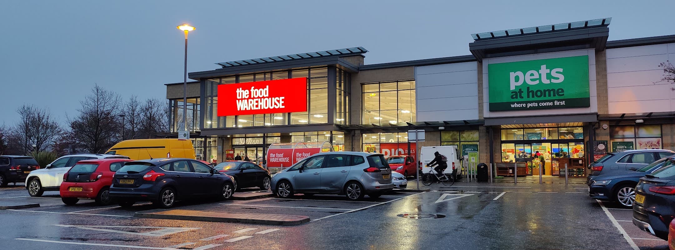 The Food Warehouse storefront with bright signage, parking lot filled with cars, and Pets at Home next door.