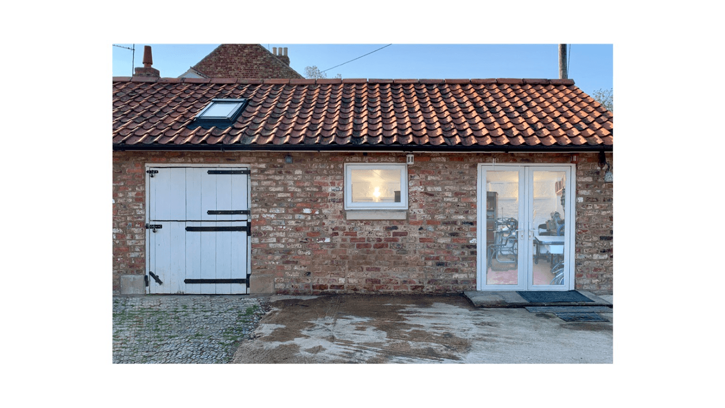 Brick building with a tiled roof, featuring a white door, a window, and visible interior through glass doors.