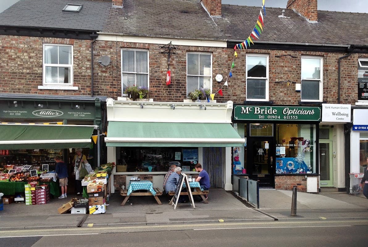 Quaint cafe with outdoor seating, green awning, and colorful bunting, next to a fruit shop and an optician's.