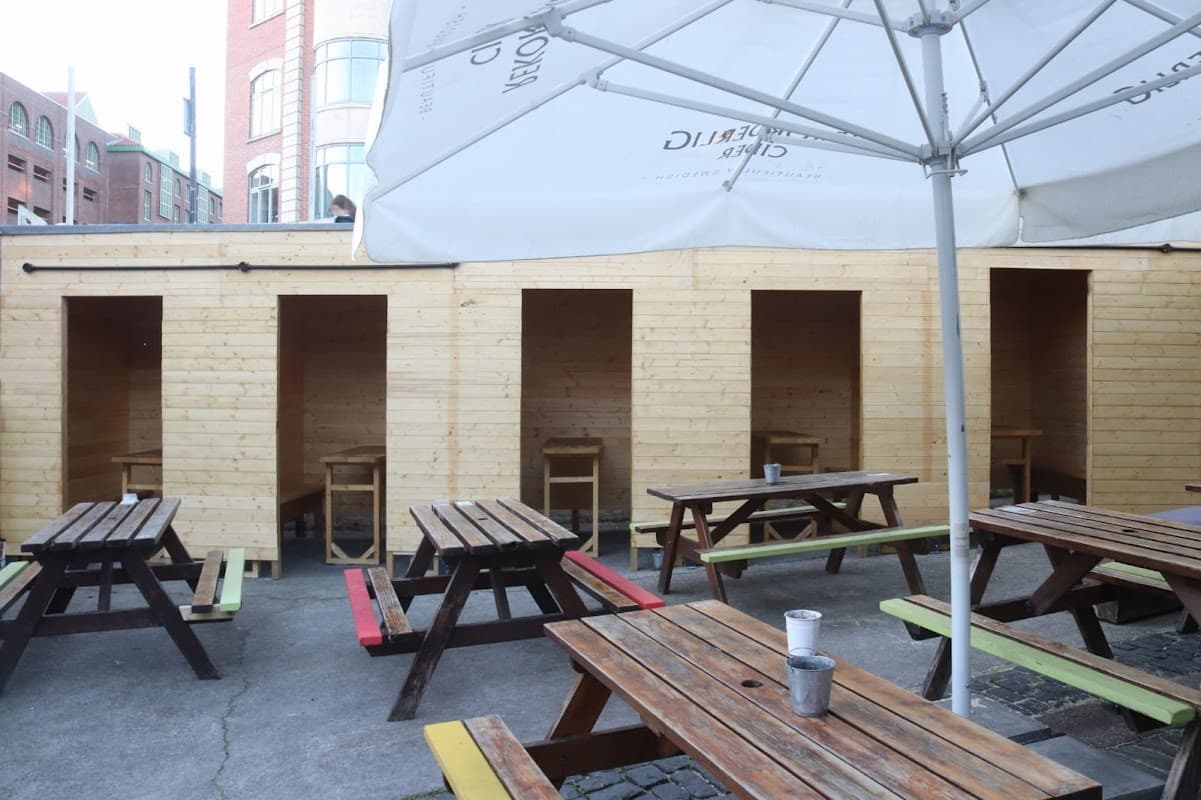 Outdoor seating area with wooden tables and chairs, sheltered by an umbrella, featuring wooden booths in the background.