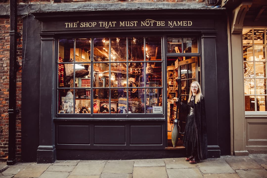 A woman in a long black dress stands outside a whimsical hobby shop with a large window displaying various colorful items.
