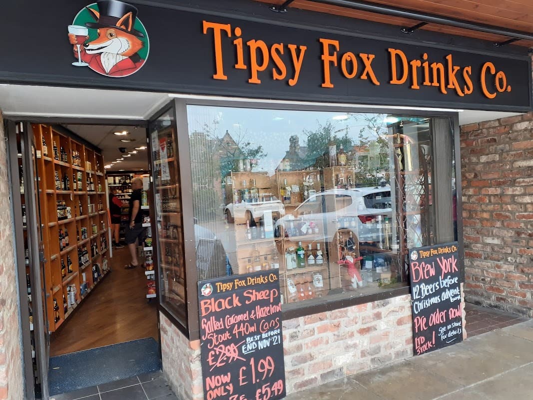 Exterior of Tipsy Fox Drinks Co with wooden shelves, signage, and promotional chalkboards displaying prices and offers.