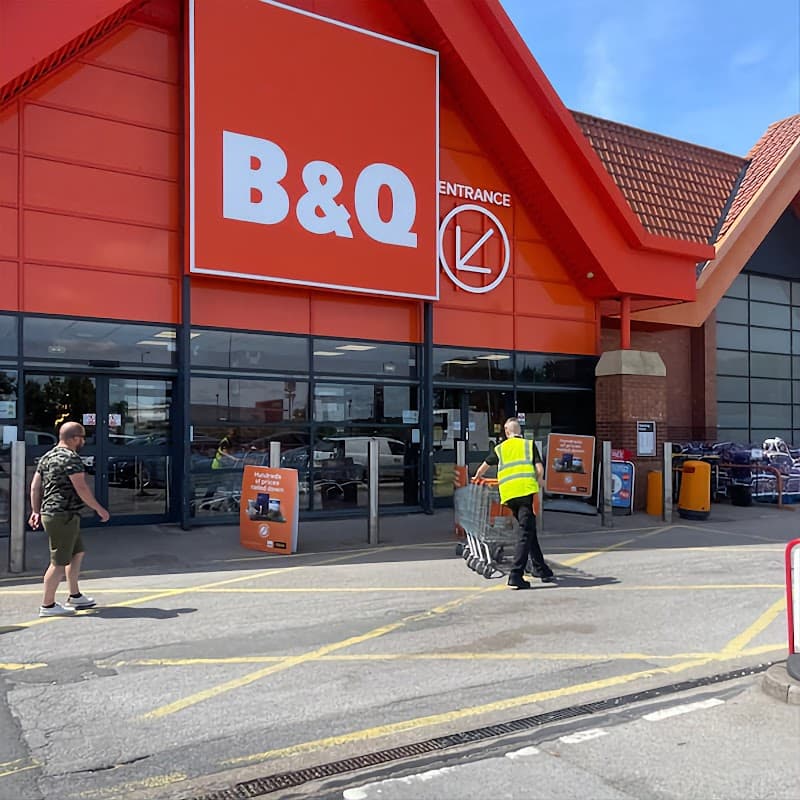 B&Q store entrance with two customers, one pushing a cart, clear blue sky, and red roof architecture.