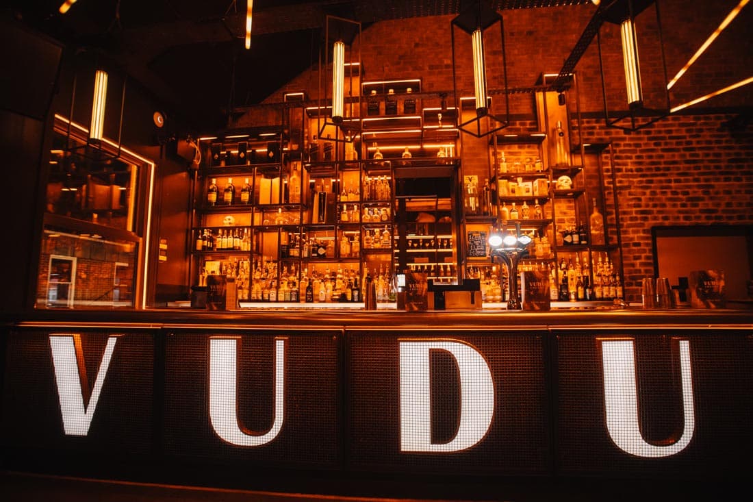 Illuminated bar with "VUDU" signage, shelves lined with bottles, and warm lighting in a stylish pub setting.
