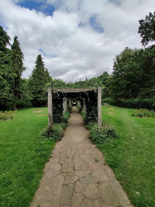 A stone pathway leads through a green park with wooden trellises covered in greenery under a cloudy sky.