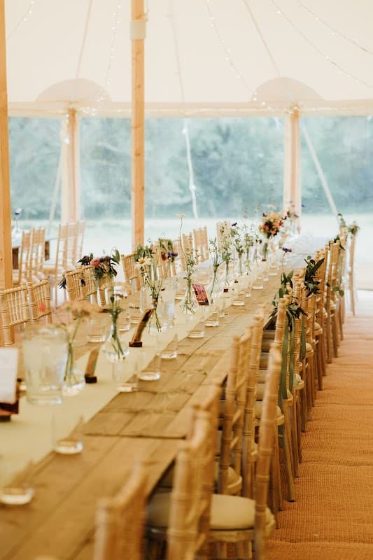 Long wooden table set with vases of flowers under a tent, surrounded by chairs and soft lighting.