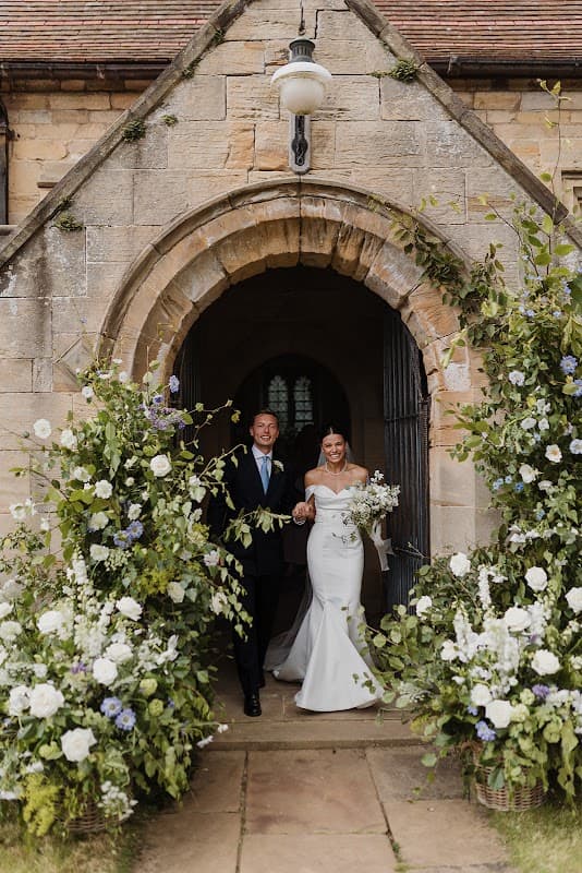 Bride and groom exit a stone archway adorned with lush floral arrangements in white and blue hues.