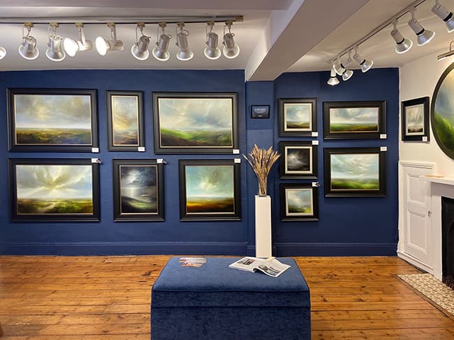 Gallery interior featuring framed landscape paintings on blue walls, wooden flooring, and a decorative plant display.